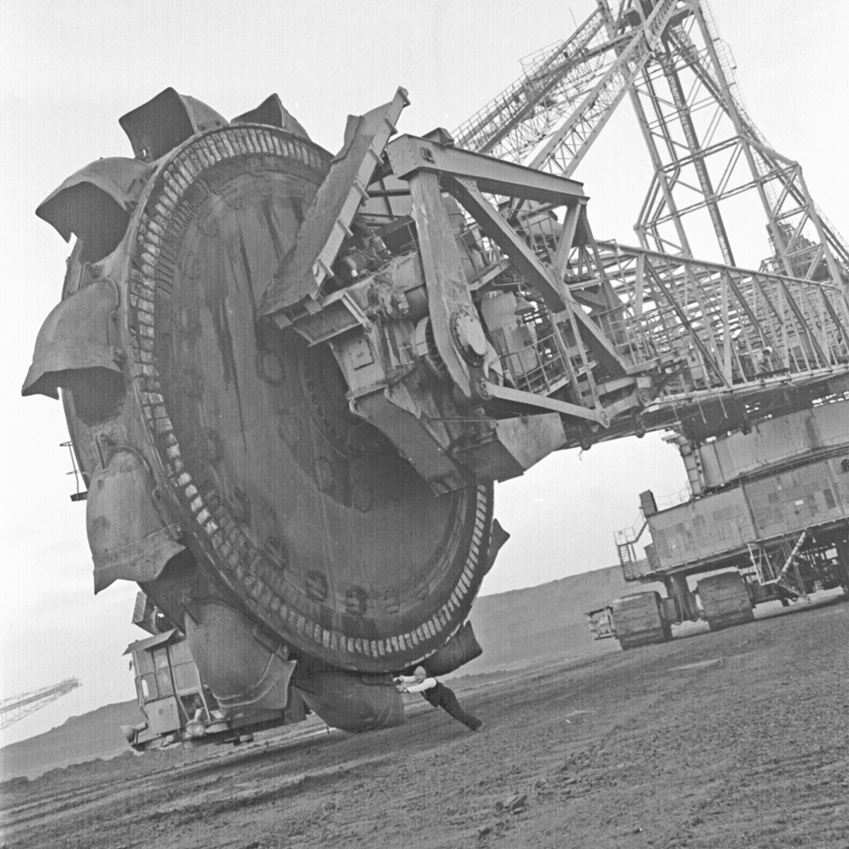 Black and white photograph of a tiny man trying to push a huge excavator bucket wheel. The man is wearing a white shirt with a black vest, black trousers and a grey wooly hat.