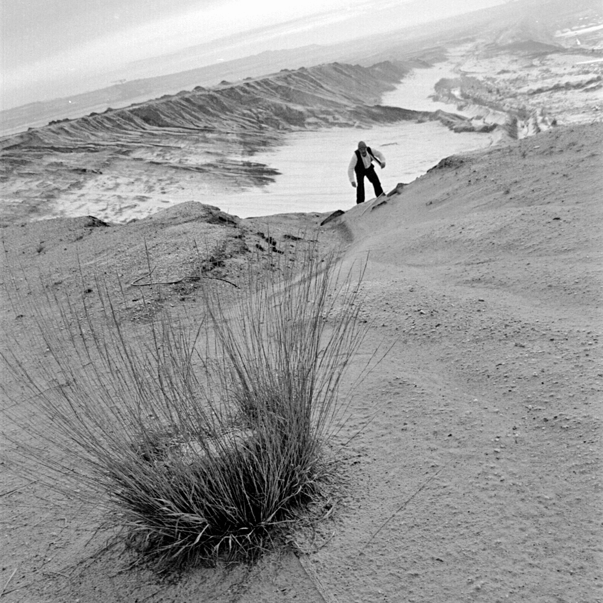 Black and white photograph of a man swaying, having reached the top of a vast empty landscape. The man is wearing a white shirt with a black vest, black trousers and a grey wooly hat.