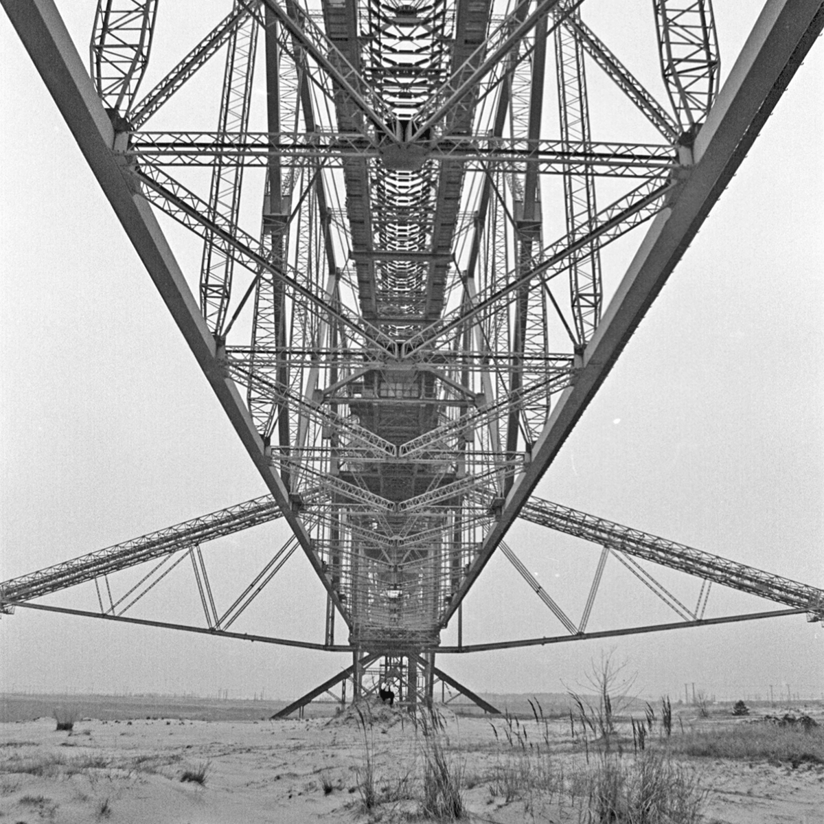 Black and white photograph of a tiny man sitting underneath a huge old metal-framed conveyor bridge. The man is wearing a white shirt with a black vest, black trousers and a grey wooly hat.