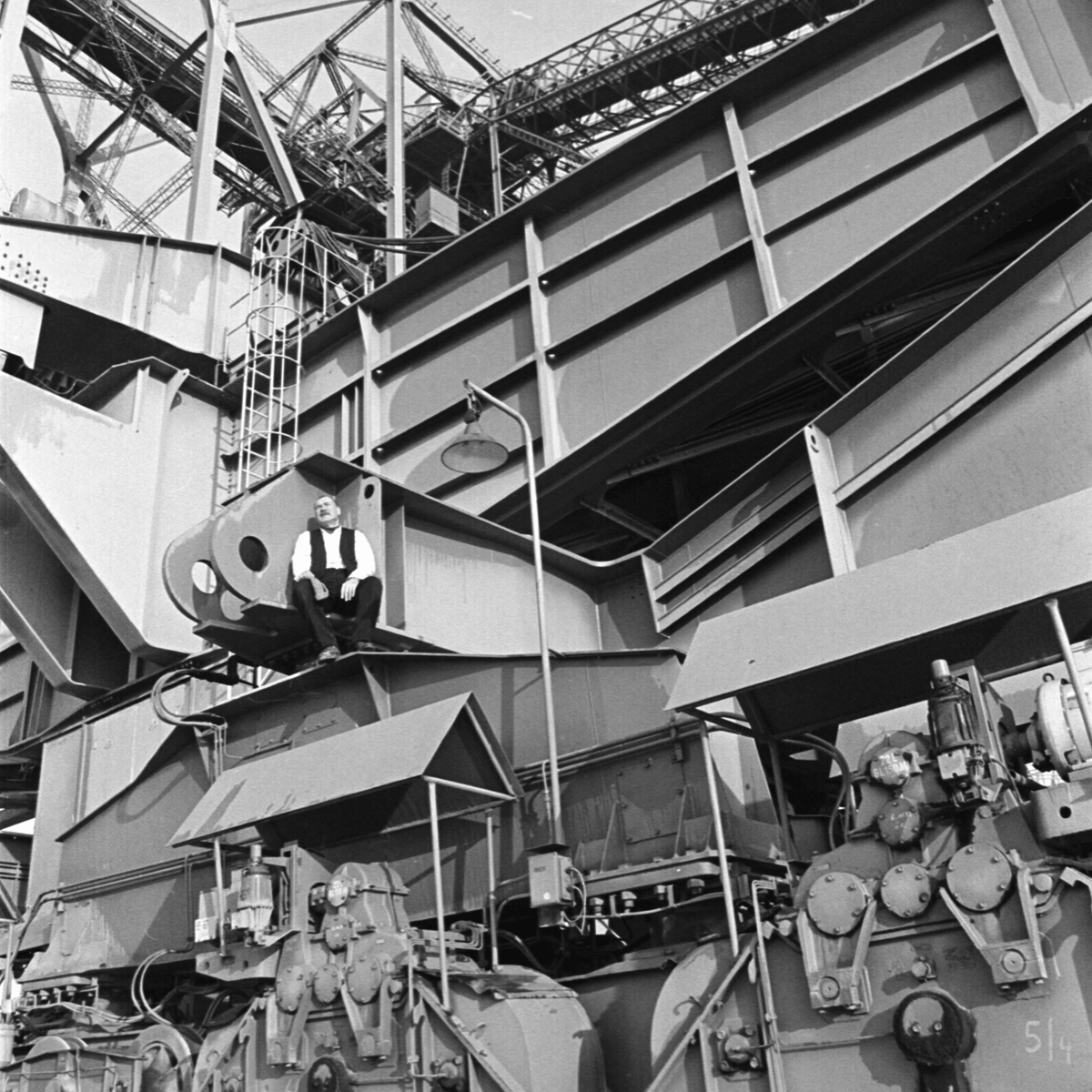 Black and white photograph of a man resting within a metal framework of a large machine. The man is wearing a white shirt with a black vest, black trousers and has a grey wooly hat in his hands.