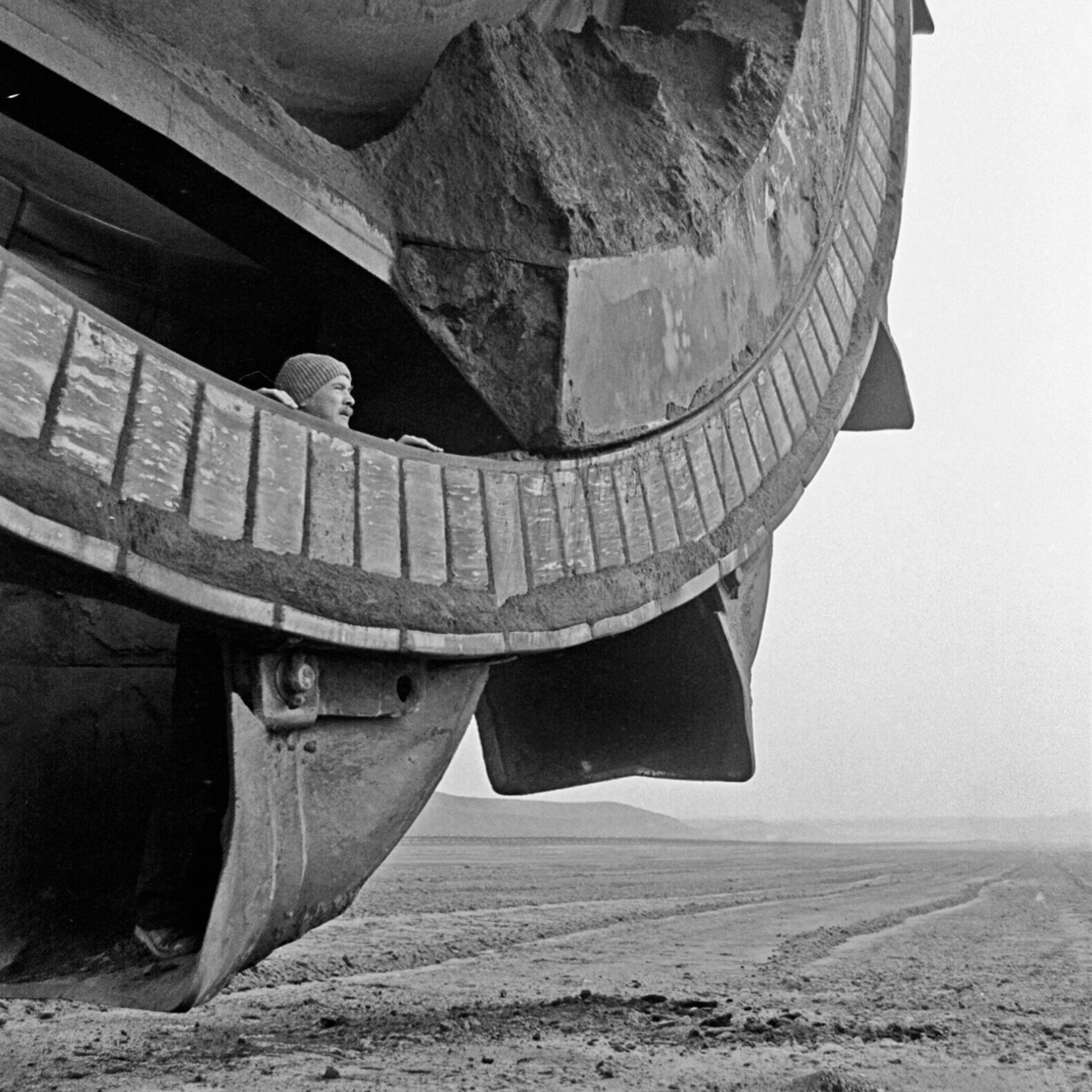 Black and white photograph of a man standing inside a bucket of a huge excavator bucket wheel. He is looking into the vast landscape.