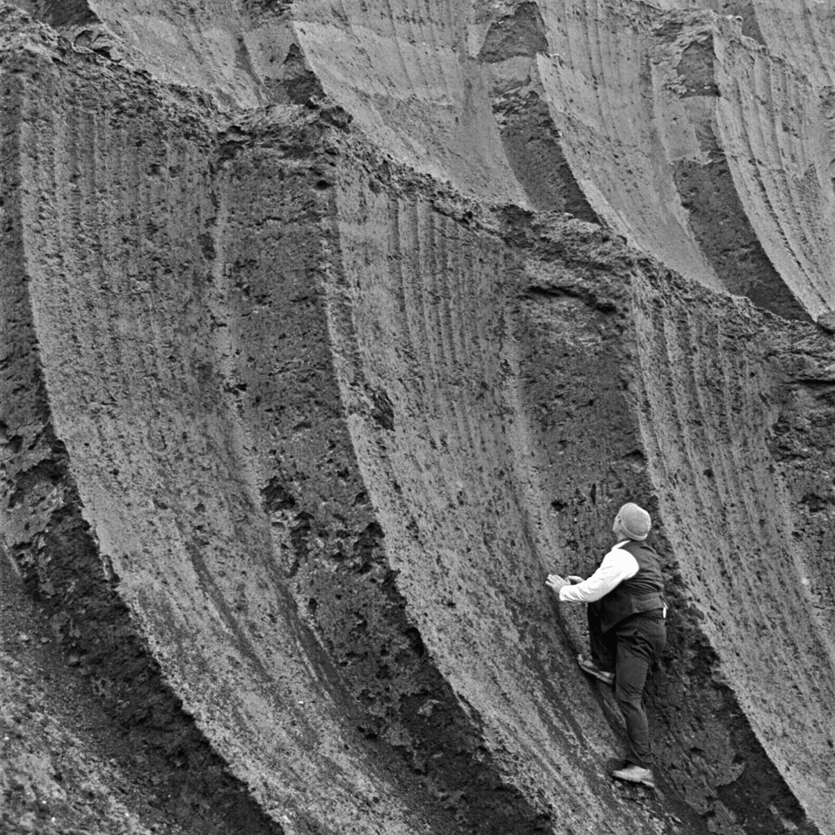 Black and white photograph of a small man trying to climb a huge wall of overburden. The man is wearing a white shirt with a black vest, black trousers and a grey wooly hat.