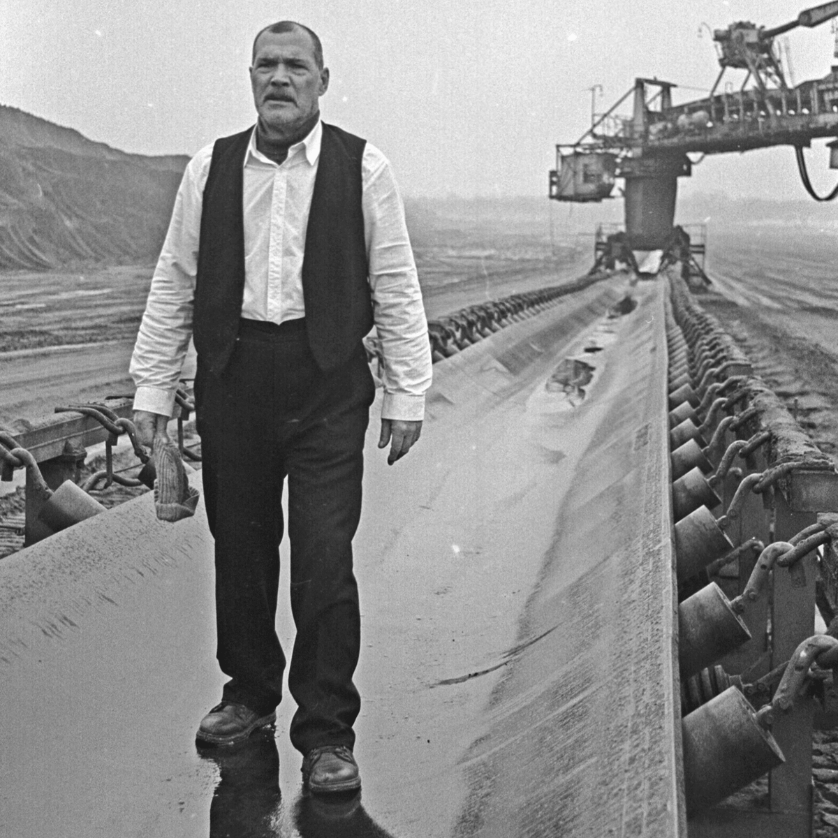 Black and white photograph of a man walking along a conveyor belt. The man is wearing a white shirt with a black vest, black trousers and has a grey wooly hat in his hand.