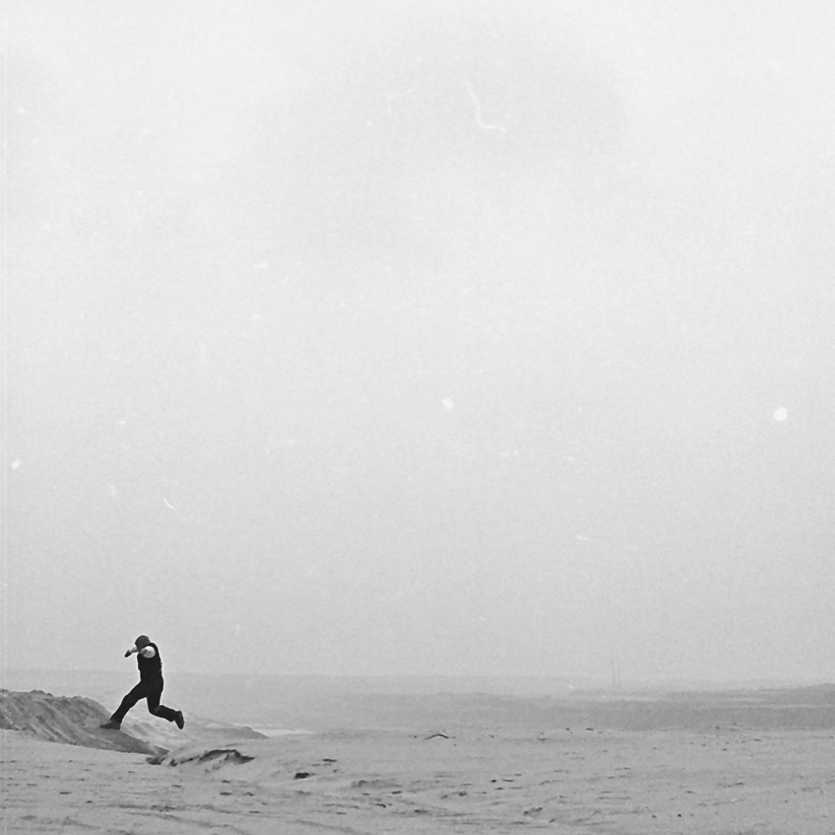 Black and white photograph of a man jumping across a rut in a vast empty landscape. The man is wearing a white shirt with a black vest, black trousers and a grey wooly hat.