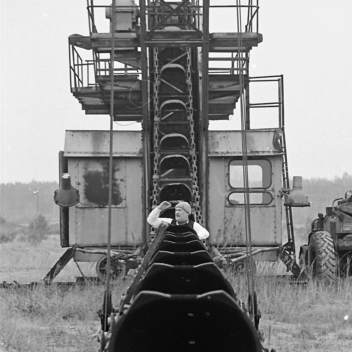 Black and white photograph of a man sitting in a small conveyor between excavator buckets. The man is wearing a white shirt with a black vest, black trousers and a grey wooly hat.