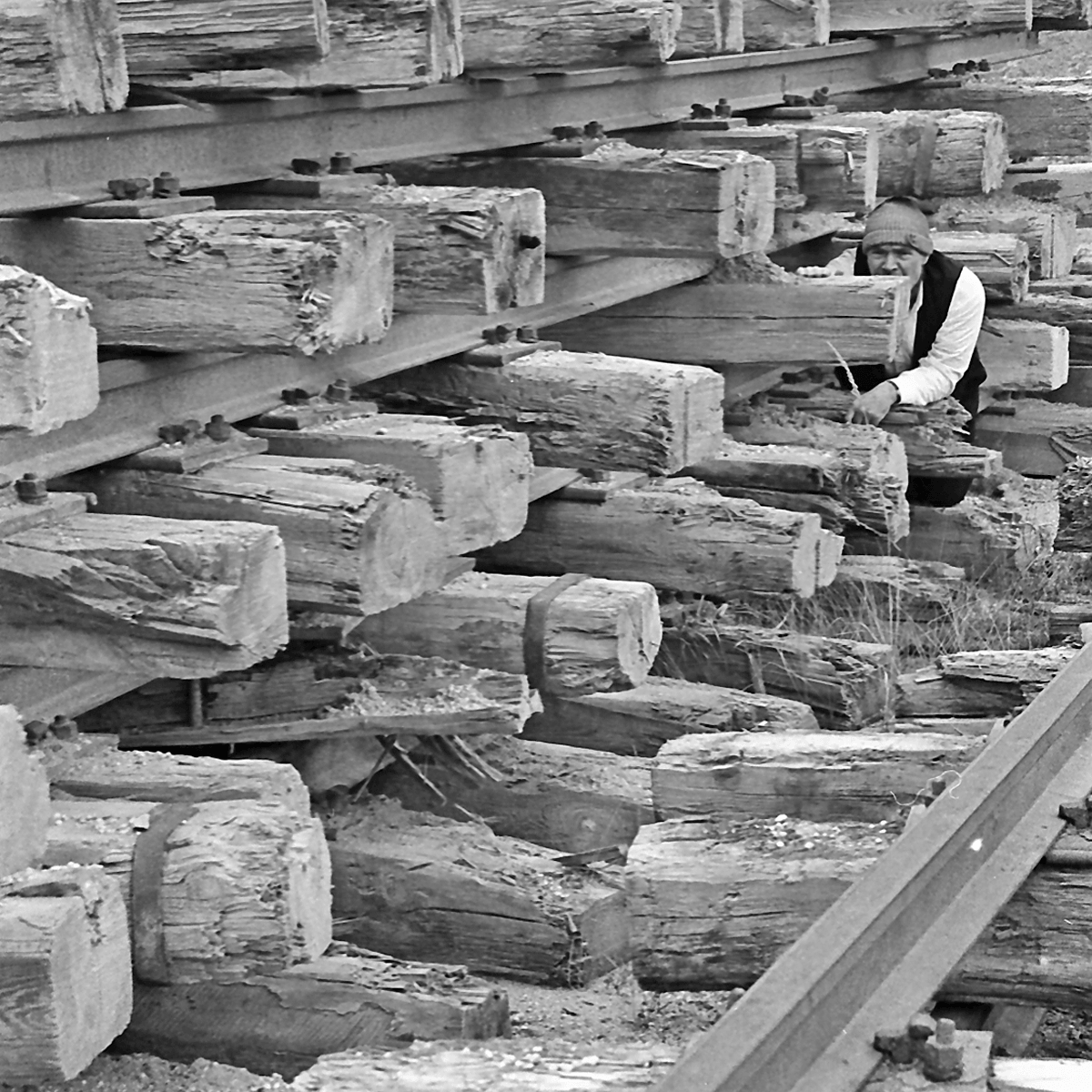 Black and white photograph of a man hiding between stacked old rails and sleepers. The man is wearing a white shirt with a black vest, black trousers and a grey wooly hat.