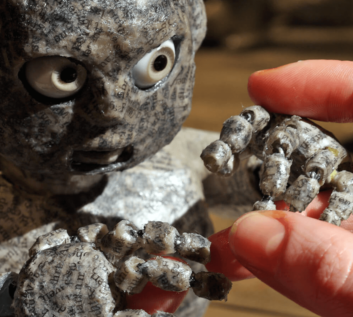 Close-up photograph of a papier-mâché puppet with glass eyes full of expression. A human hand is carefully manipulating the puppet's hands